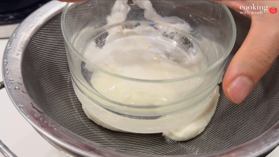 Glass bowl pressing down on cheesecloth bundle in a fine mesh sieve, draining whey from hung curd.
