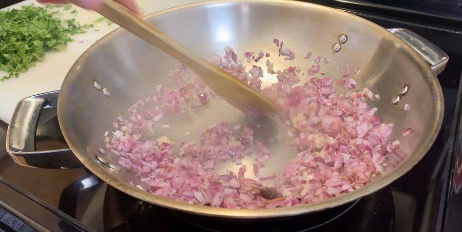 Diced pink onions sautéing in a large pan with a wooden spoon, partially cooked and translucent.