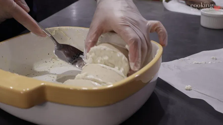 Hands transferring rolled gluten-free strukli dough pieces into a buttered yellow and white baking dish.