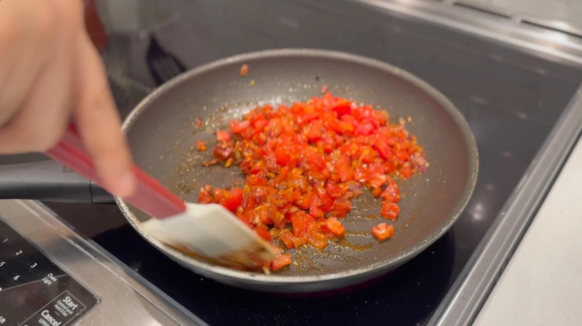 Diced red bell peppers and onions being sautéed in a gray pan with a wooden spoon on a stovetop.