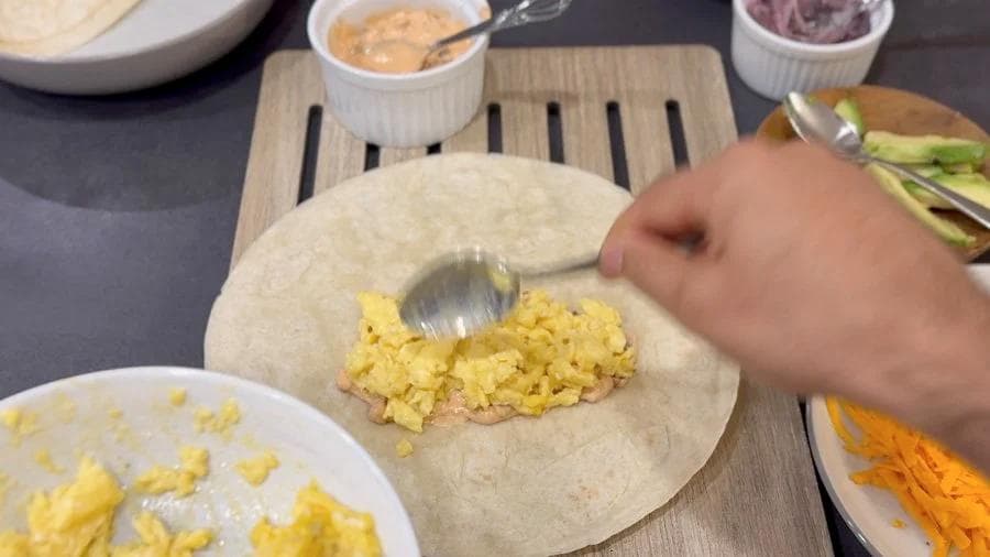 Hands folding scrambled eggs into a flour tortilla on a wooden board while preparing a breakfast burrito.