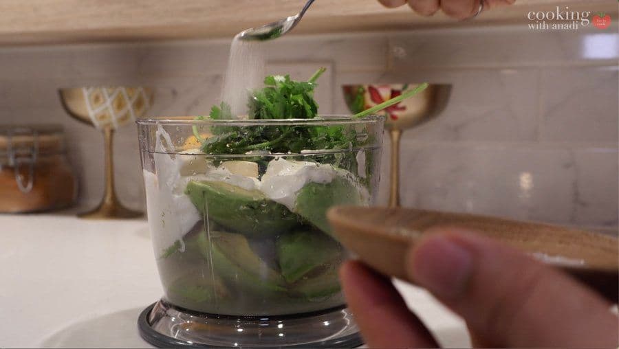 Food processor blending avocado, lime, cilantro, and other ingredients into creamy sauce.