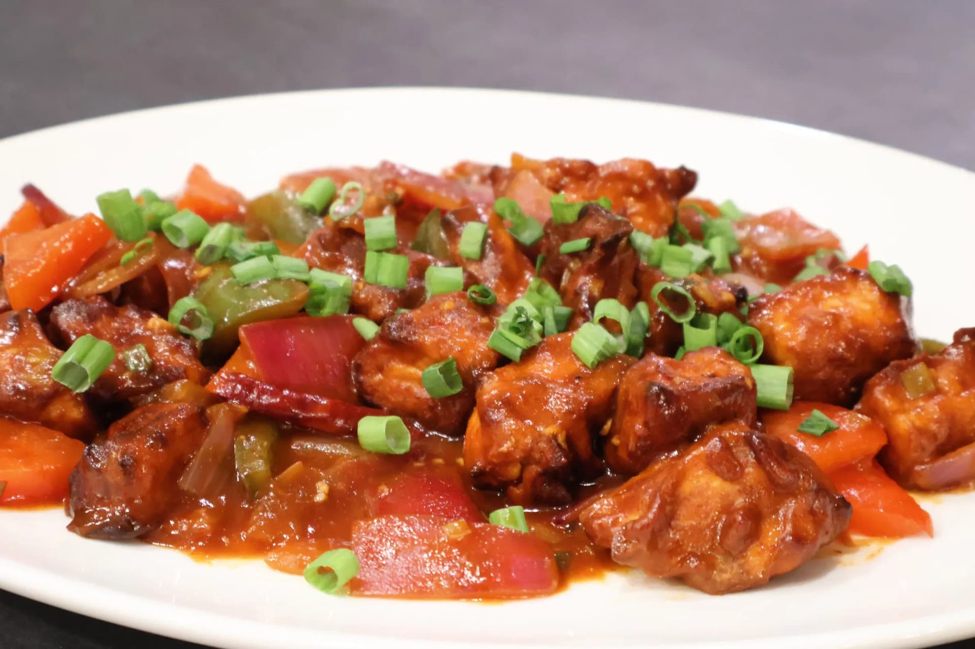 Chilli paneer stir-fry with red and green bell peppers in spicy sauce, garnished with sliced green onions on a white plate.