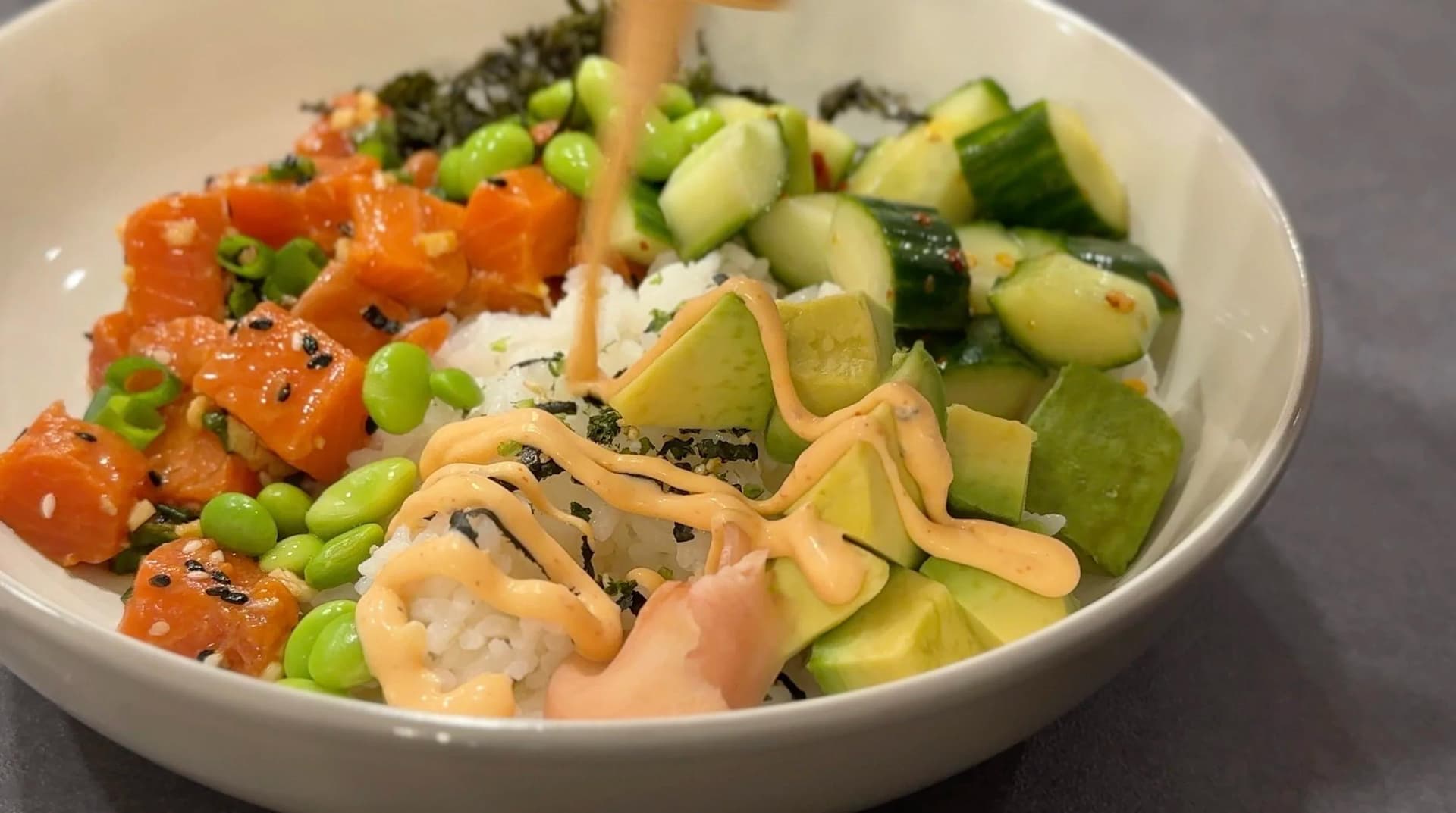 Assembled salmon poke bowl with rice, salmon cubes, edamame, cucumber, avocado, nori strips, and furikake in a white bowl.