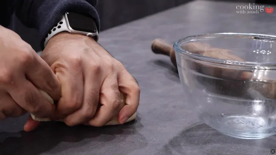 Hand kneading dough on dark surface with bowl of water nearby for Delhi-style bhature preparation.