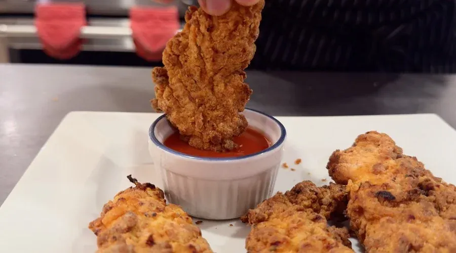 Crispy spicy chicken tender being dipped into red dipping sauce, with more tenders on a white plate beside it.