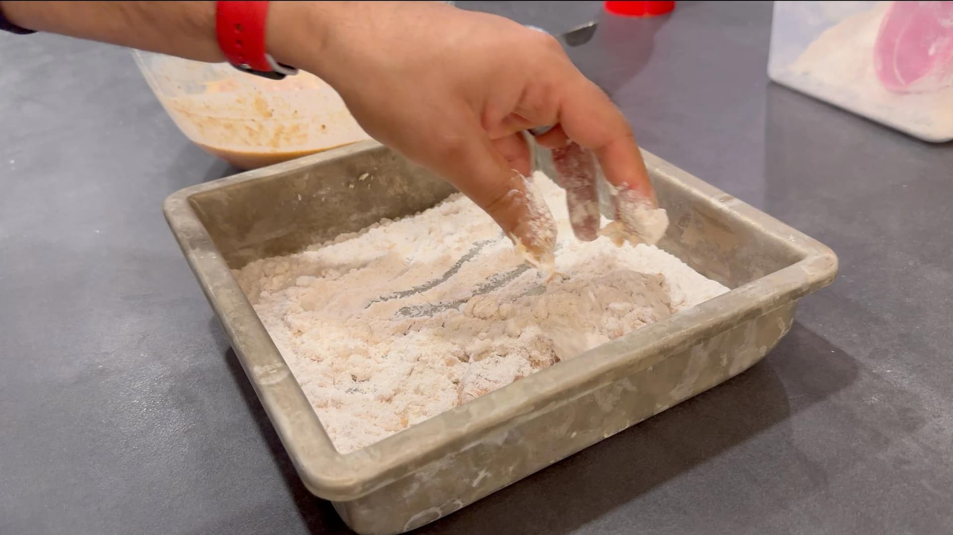 Hand coating chicken tenders in seasoned flour mixture in a baking pan during breading preparation.