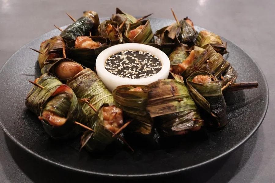 Pandan leaf-wrapped chicken pieces arranged on a black plate around a dipping sauce with sesame seeds.