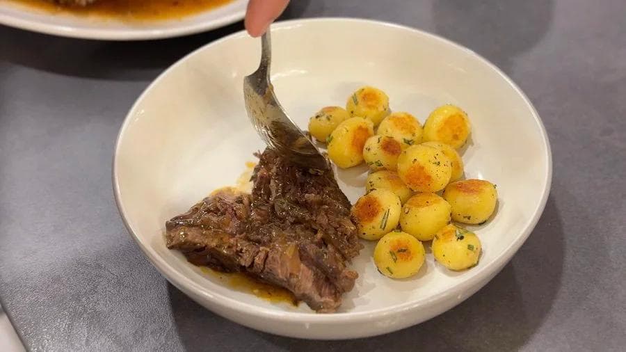 Plated pot roast with herb-roasted potatoes, fork lifting tender meat portion from white bowl.