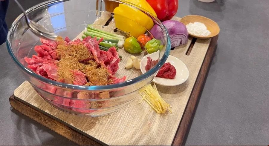 Sliced flank steak coated with Jamaican jerk seasoning and spices in a glass bowl, surrounded by fresh ingredients including peppers, onions, and garlic.