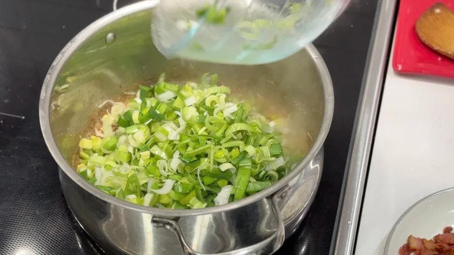 Chopped green onions and leeks being added to a pot with browned chicken and bacon fat for sautéing.