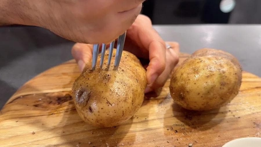 Fork pricking raw potatoes on wooden board after seasoning with oil, salt, and pepper.