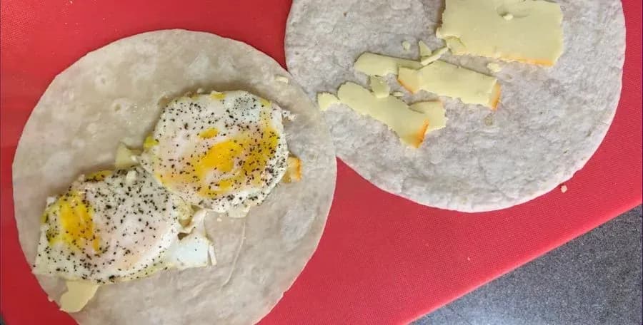 Two tortillas with assembled breakfast wraps showing eggs, cheese, and black pepper seasoning on a red surface.