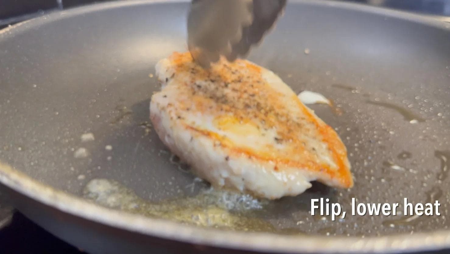 Seared chicken breast being flipped in a pan with garlic oil, showing golden-brown crust on one side.