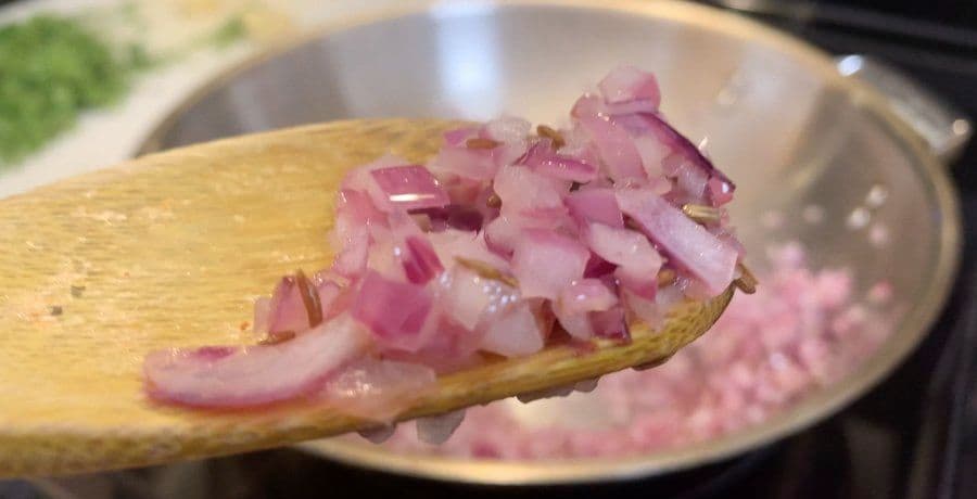 Diced pink onions on a wooden spoon being added to a pan during the sautéing step of paneer preparation.