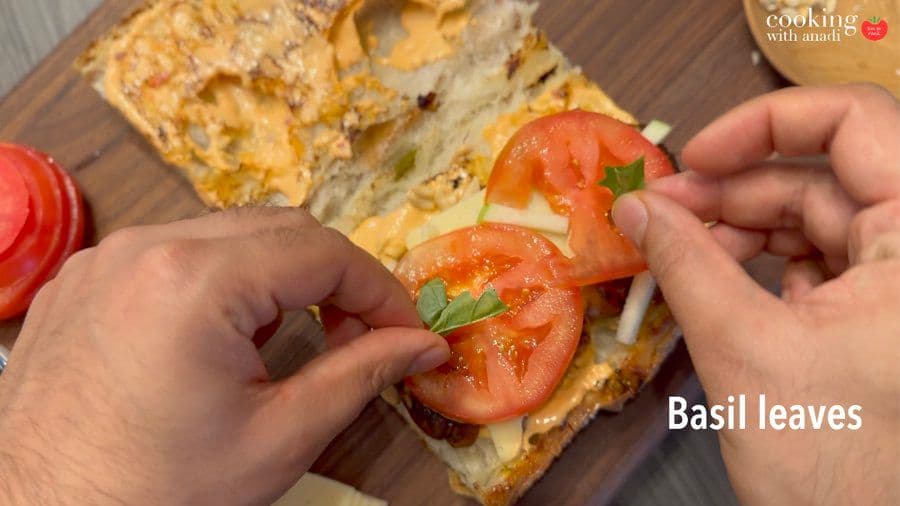 Hands placing fresh basil leaves and tomato slices on toasted baguette with curry mayo and other fillings.
