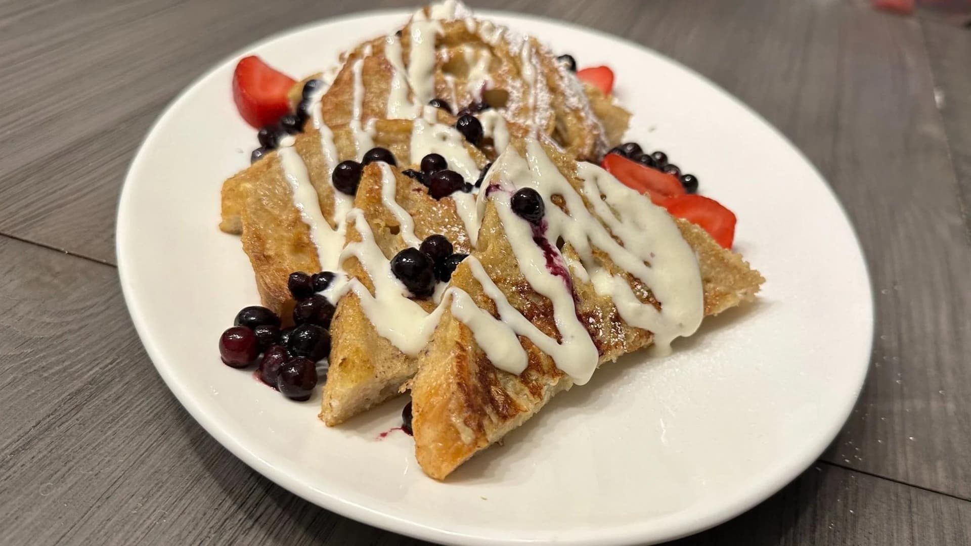 Plated French toast triangles drizzled with white icing, topped with dark berries and fresh strawberry slices on a white plate.