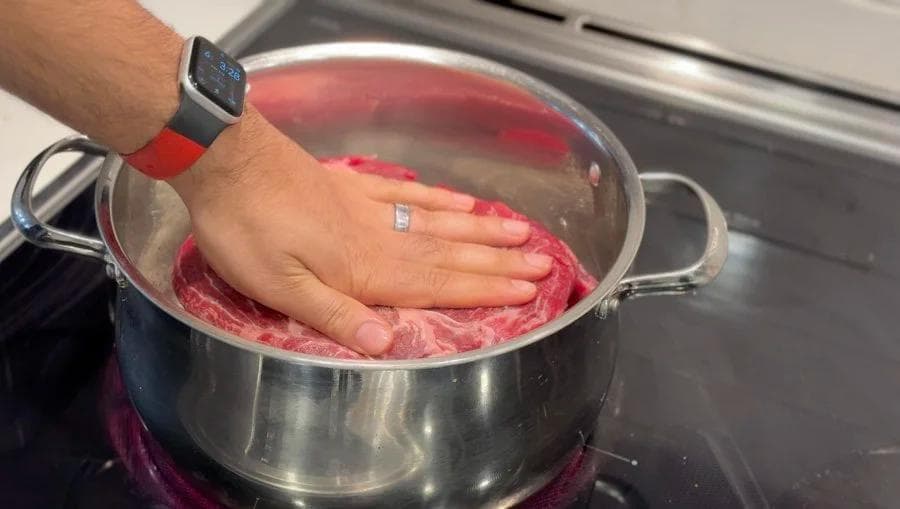 Hand pressing seared beef into a pot of red liquid during pot roast preparation.