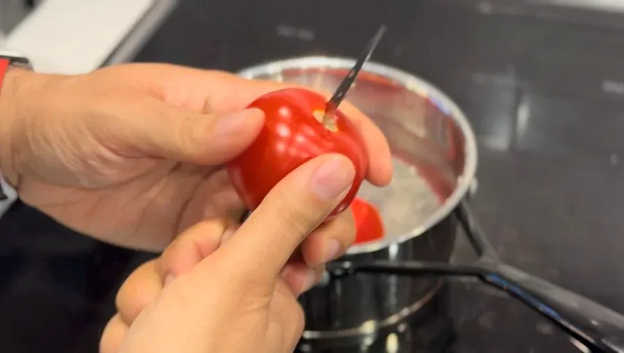 Hand holding a red tomato with a fork over a saucepan of water containing tomato halves and dried red chillies.