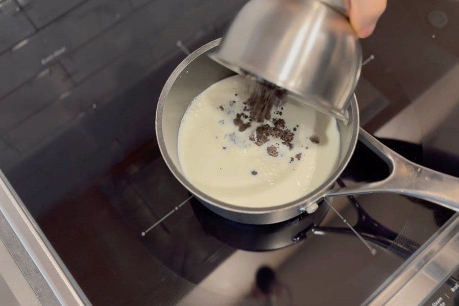 Pouring crushed Oreo cookies into steaming milk in a saucepan on a stovetop.
