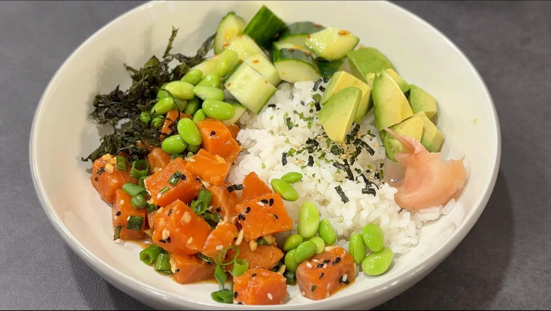 Assembled salmon poke bowl with white rice, cubed salmon, cucumber, edamame, avocado, seaweed, and furikake in a white bowl.