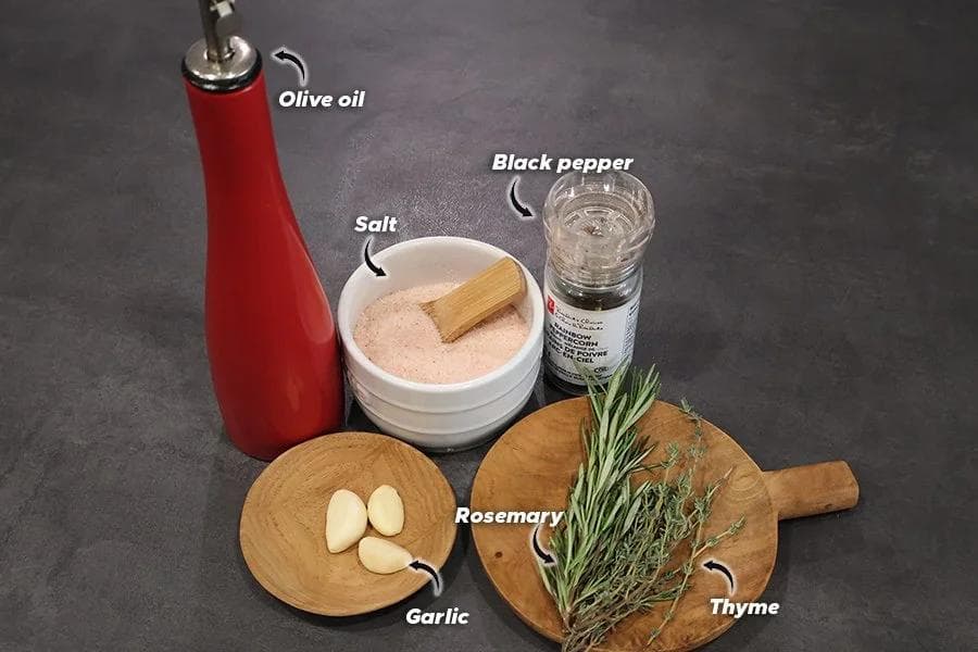 Six garlic-herb oil ingredients on a gray surface: red olive oil bottle, pink salt bowl, black pepper grinder, garlic cloves, rosemary, and thyme.