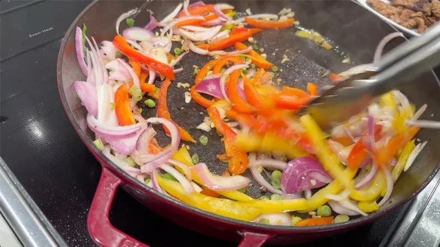 Sautéing sliced bell peppers, onions, and ginger-garlic in a burgundy skillet during Jamaican pepper steak preparation.