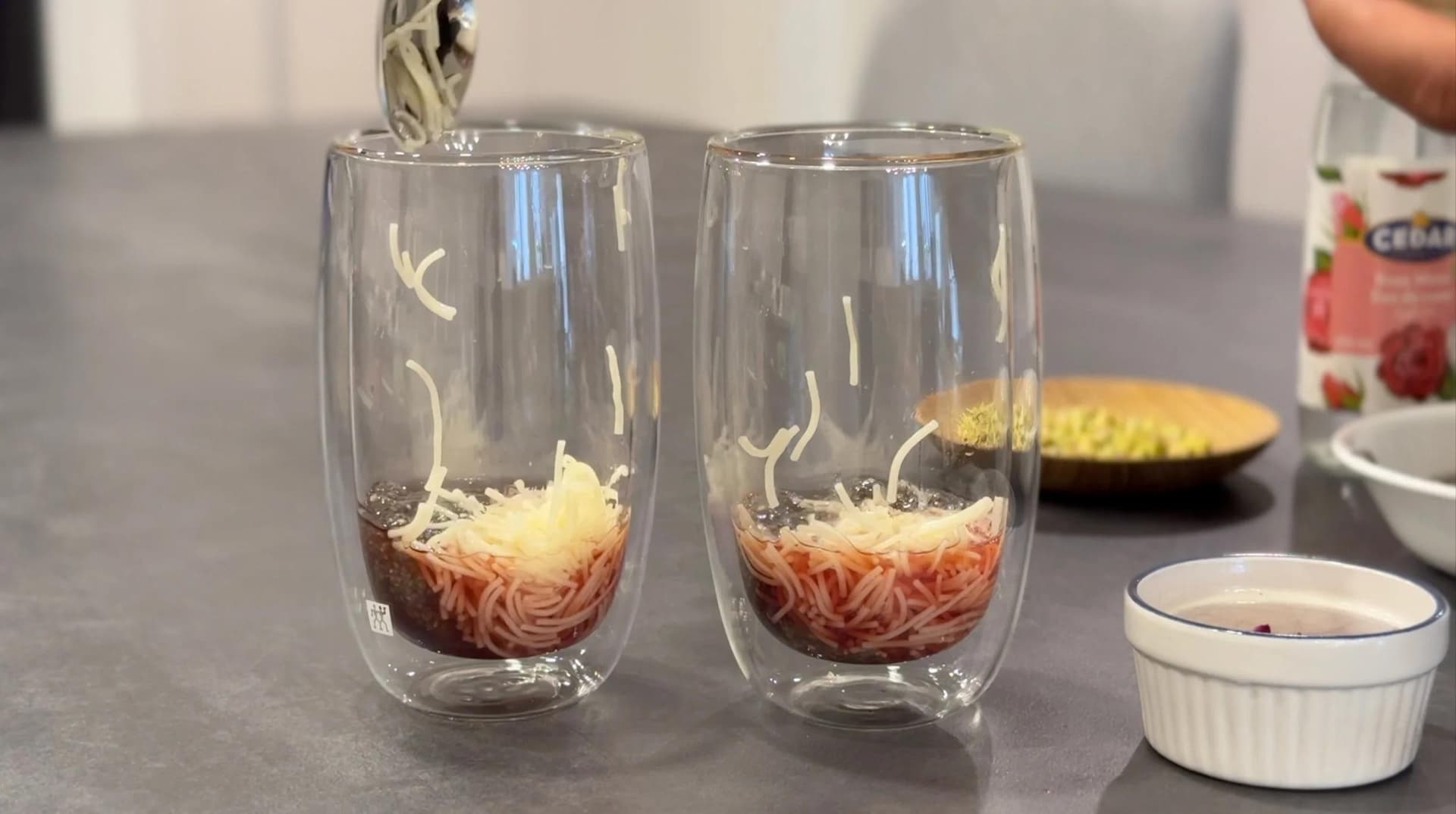 Two glasses layered with vermicelli and chia seeds, with milk being poured into one glass to make falooda.
