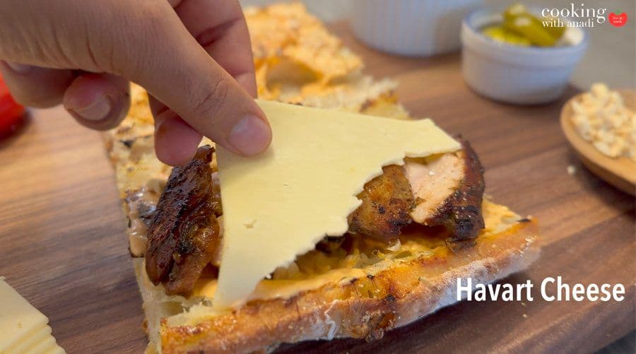 Hand placing havarti cheese slice on Thai chicken sandwich with grilled chicken and peanuts on toasted baguette.