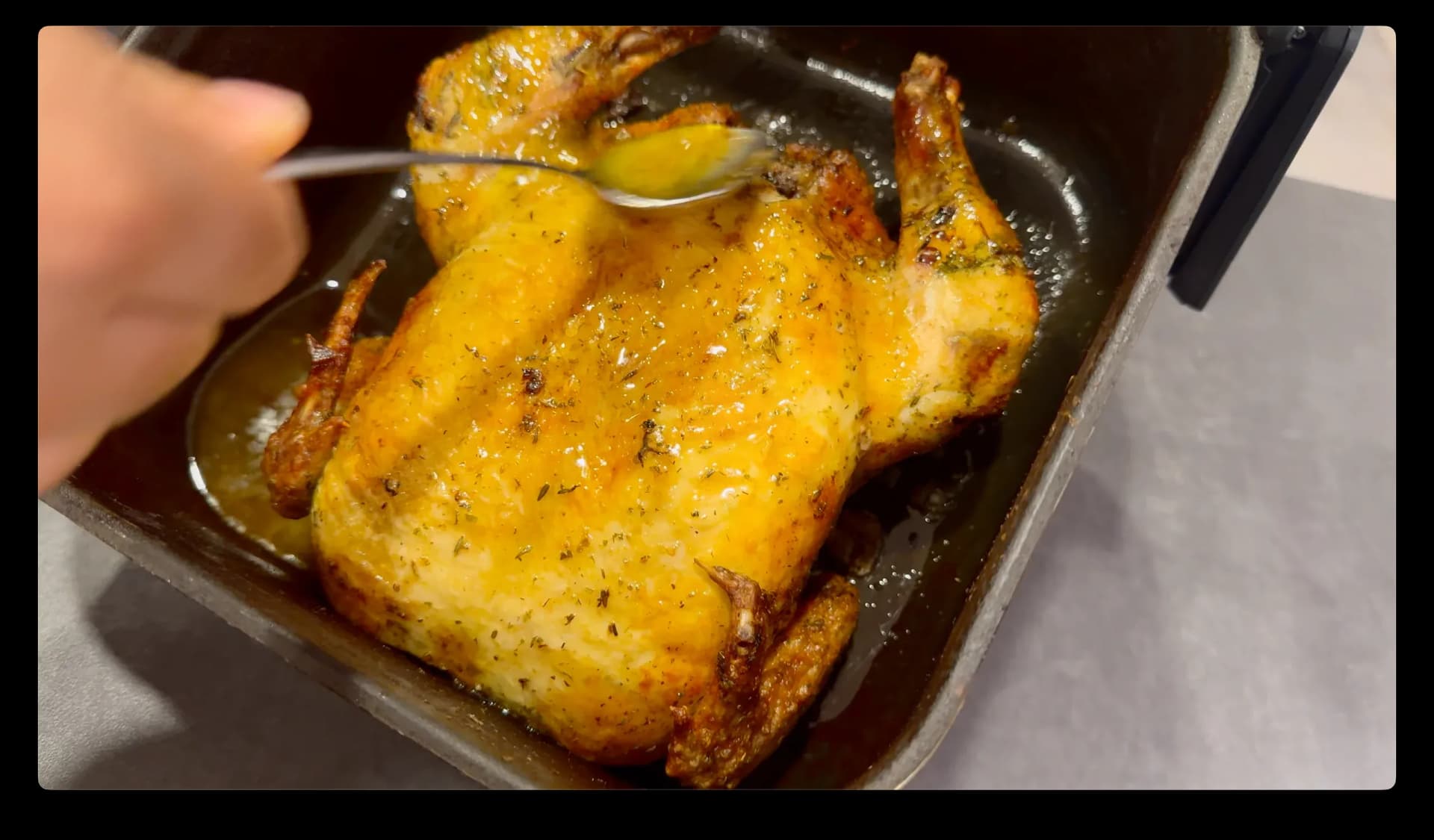 Golden-brown whole chicken in air fryer basket being basted with pan juices using a spoon.