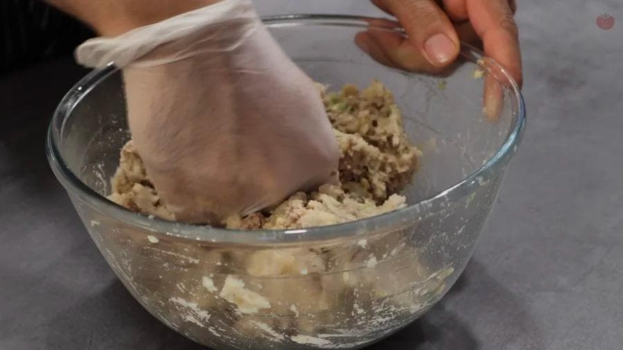 Hands mixing ground meat and potato mixture in a glass bowl with a gloved hand.