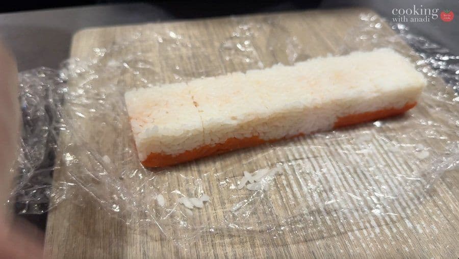 Rectangular block of oshizushi pressed sushi with white topping and red layer, partially cut into pieces on plastic wrap.