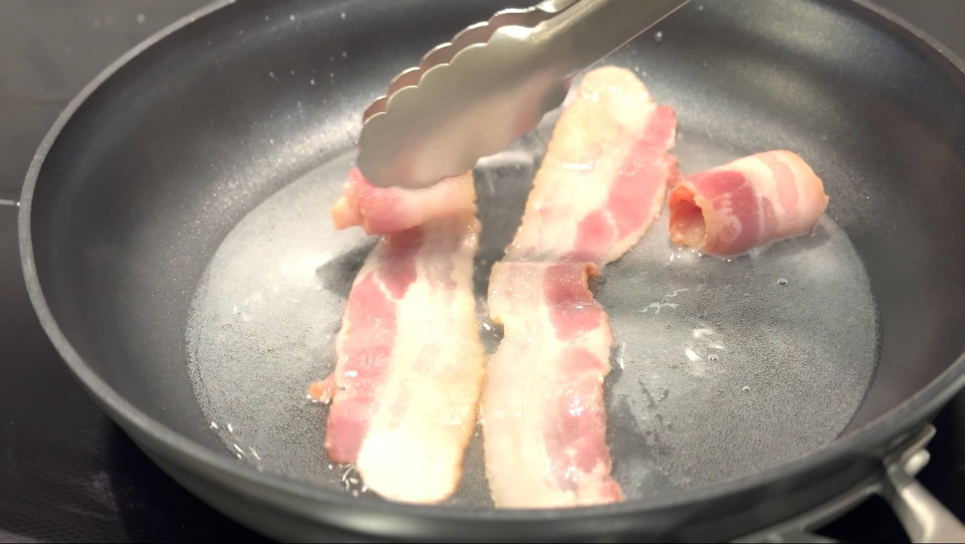 Bacon strips cooking in a dark skillet with water, being turned with tongs as fat renders.
