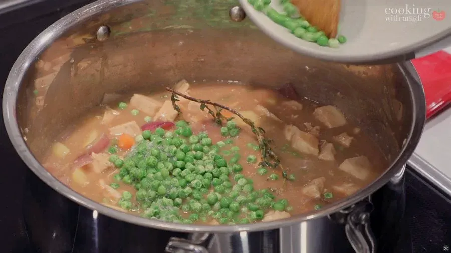 Green peas and herb sprigs added to simmering Guinness chicken stew in a red pot.