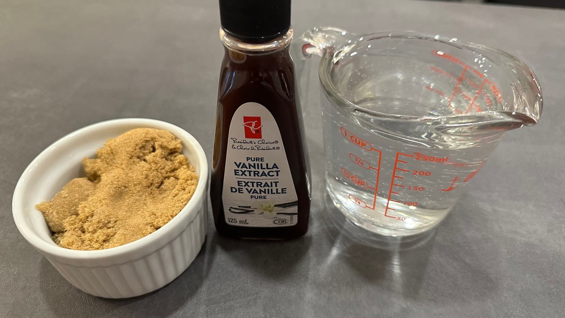 Three ingredients for homemade brown sugar syrup: brown sugar in white bowl, vanilla extract bottle, and water in measuring cup.