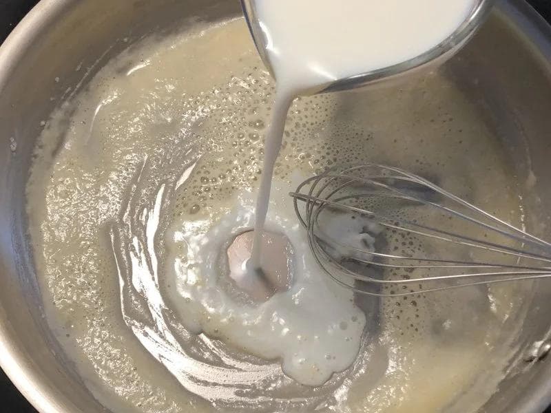 Milk being poured into a roux while whisking to create bechamel sauce in a metal pot.