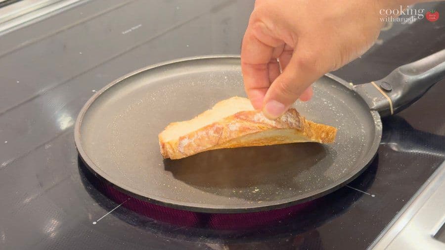 Hand placing toasted bread slice with tuna salad onto a non-stick pan during sandwich preparation.