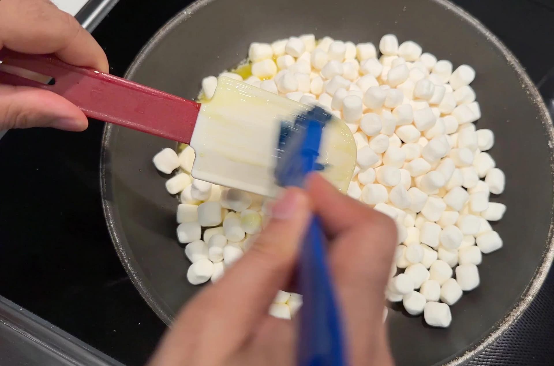Melting butter and marshmallows in a pan with a spatula and spoon during preparation of Rice Krispies treats.