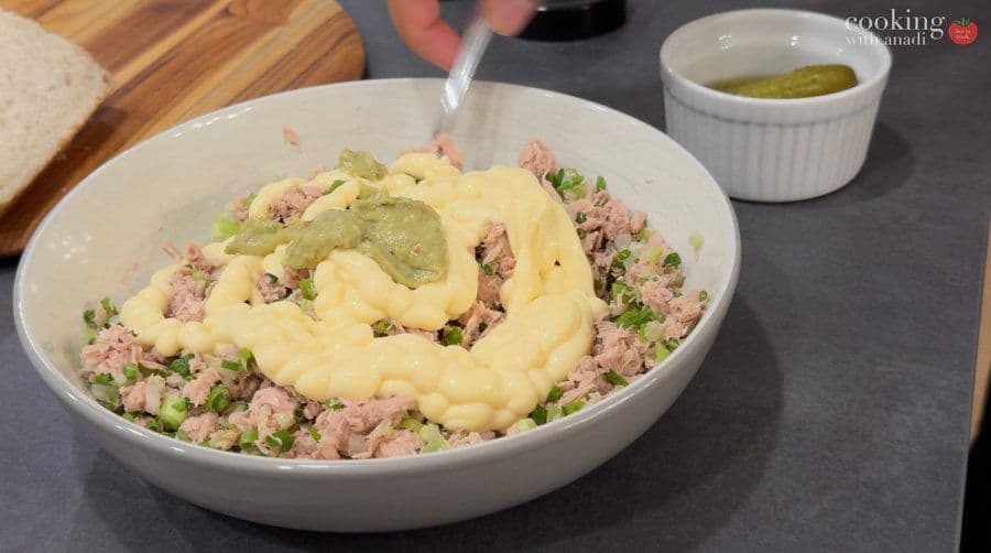 Tuna salad mixture in a white bowl with mayonnaise being stirred in, garnished with green onions and celery.