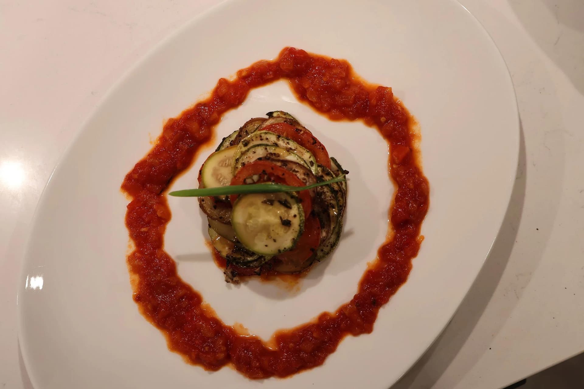 Confit byaldi ratatouille stack of layered zucchini and tomato slices, surrounded by swirled tomato sauce on a white plate.