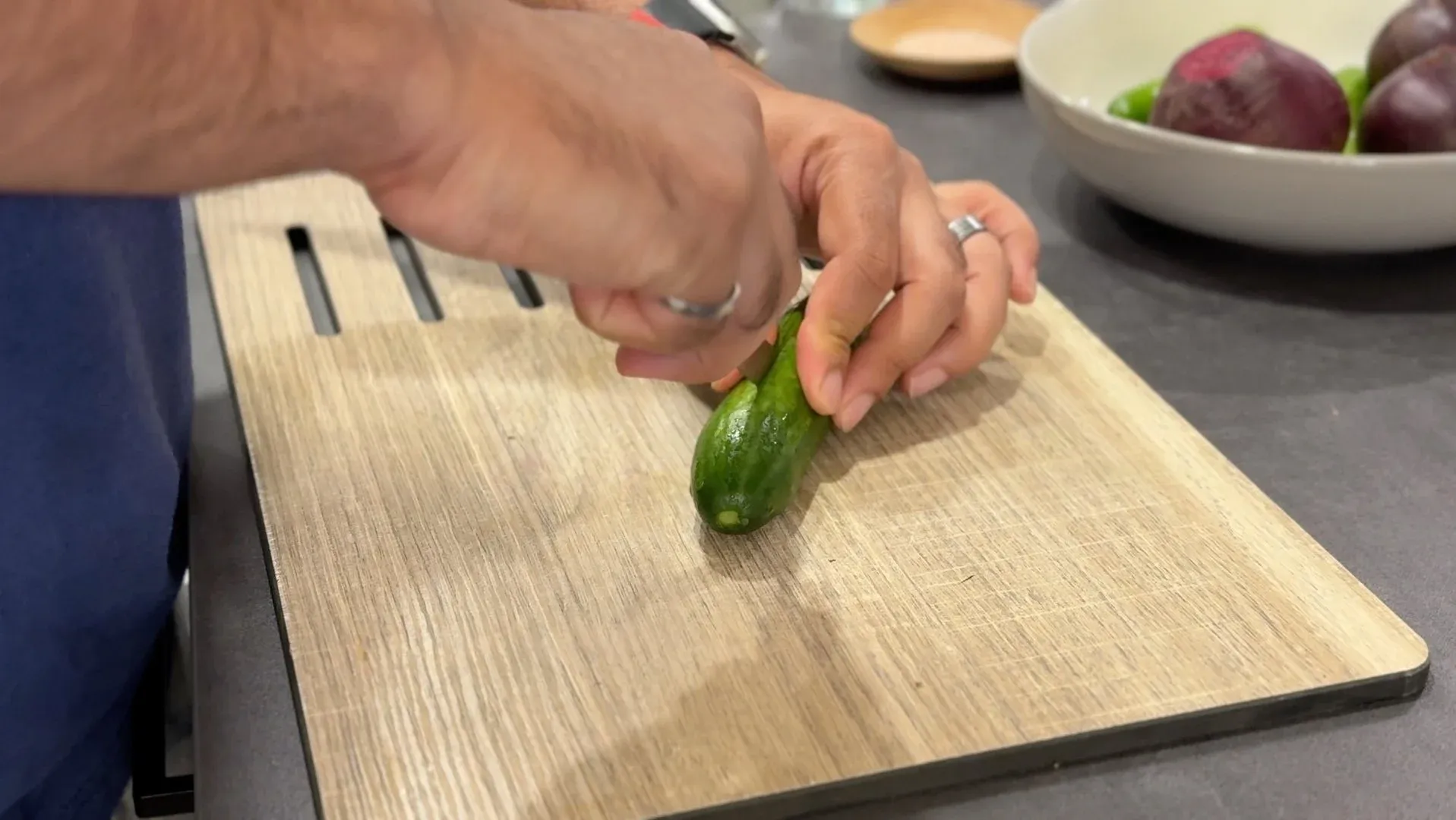 Hands cutting a baby cucumber lengthwise into quarters on a wooden cutting board.