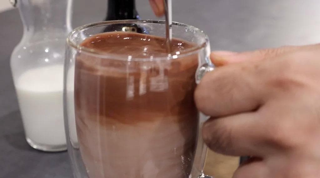Hand holding glass of creamy Baileys hot chocolate with milk pitcher in background.