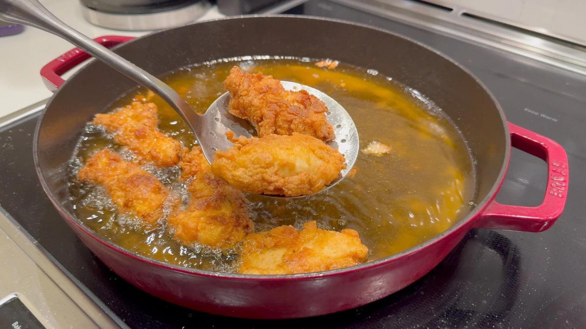 Golden fried chicken tenders cooking in hot oil in a red Dutch oven, one piece being lifted with a slotted spoon.