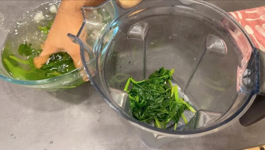 Blended spinach puree being transferred from a glass bowl into a food processor for palak paratha dough preparation.