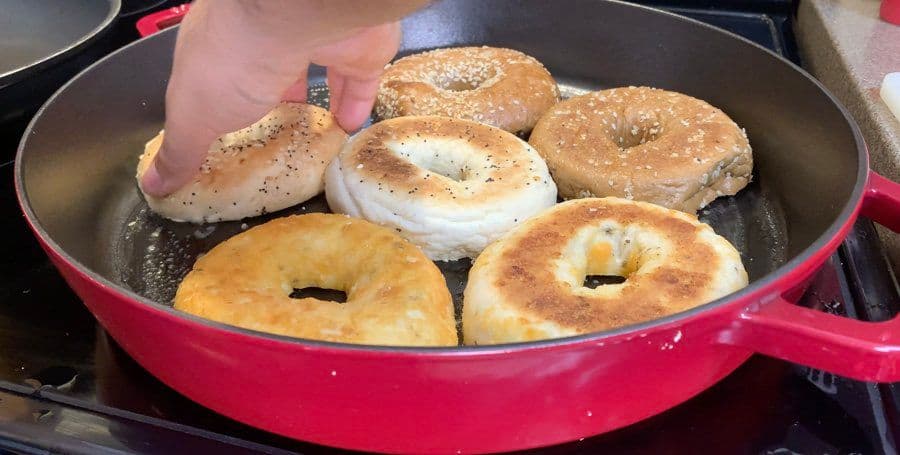 Bagels toasting in a red skillet with butter, some topped with cream cheese and seasonings.