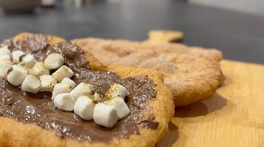 Beaver tail pastry topped with chocolate spread, cinnamon sugar, and white marshmallows on a wooden board.