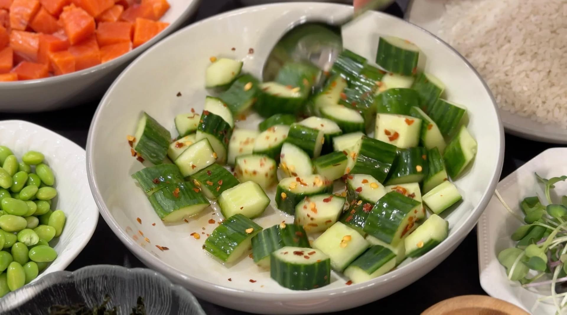 Diced cucumber mixed with chili flakes and sesame oil in a white bowl, ready to be refrigerated.