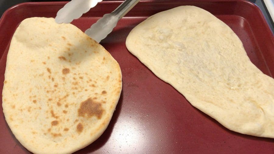 Two cooked naan breads on a red tray, one with light brown spots, being lifted with tongs.