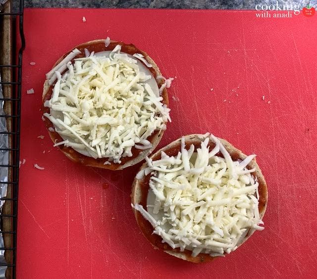 Two bagel halves topped with shredded mozzarella and provolone cheese on a red cutting board, ready for broiling.