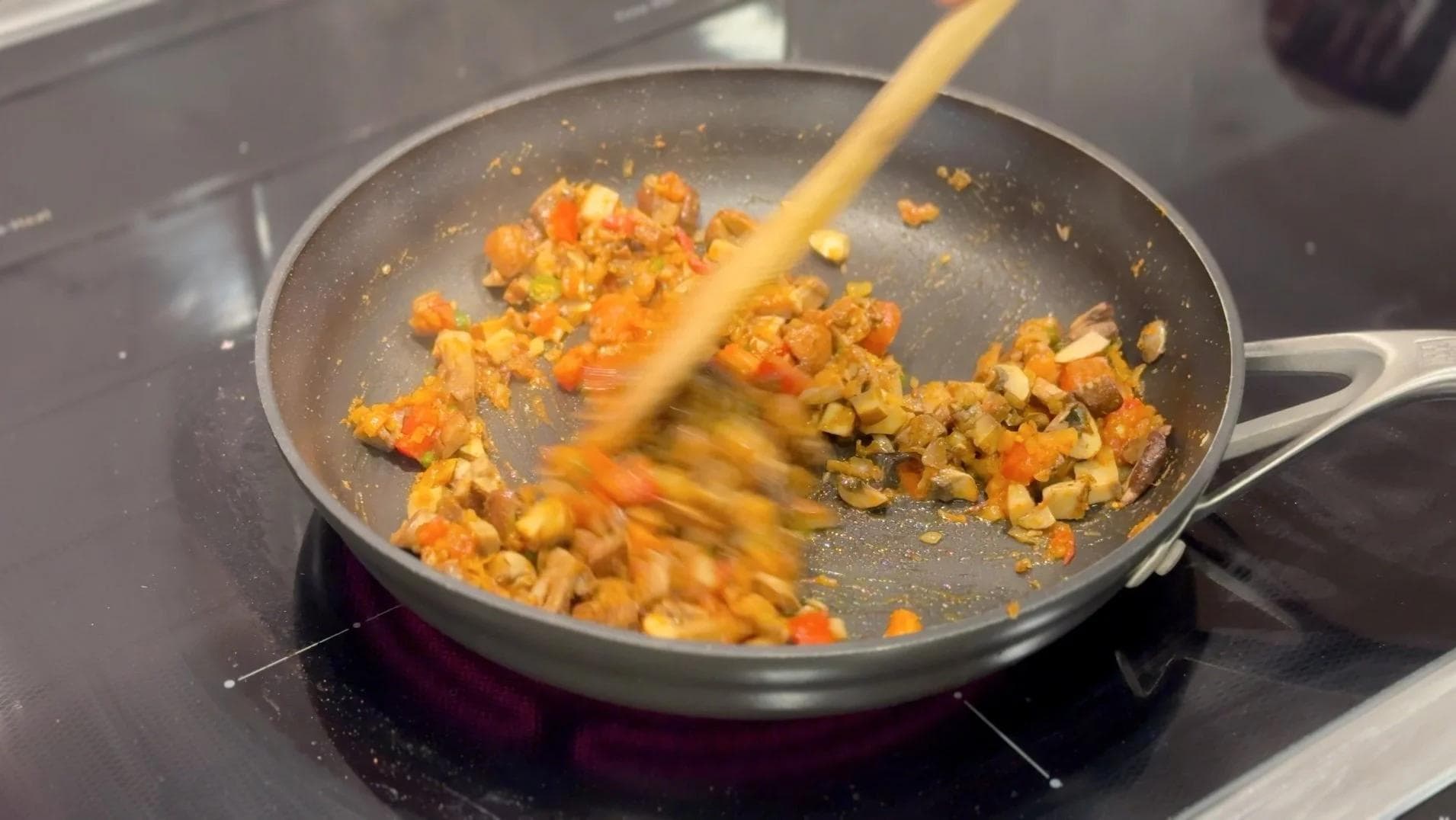 Wooden spatula stirring cooked diced mushrooms, tomatoes, and peas in a gray skillet on a stovetop.